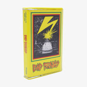 Photo of a cassette case against a white background. The album artwork on the case is yellow and on the left side going down the image is a sideways lettering that says Bad Brains. The letters are red and outlined in yellow, and then outlined again in green. On the right side of the artwork is a rectangle with the same red, yellow, green outline. Inside the outlines is a black and white drawn image of the Capitol Building being struck by yellow lightning. 